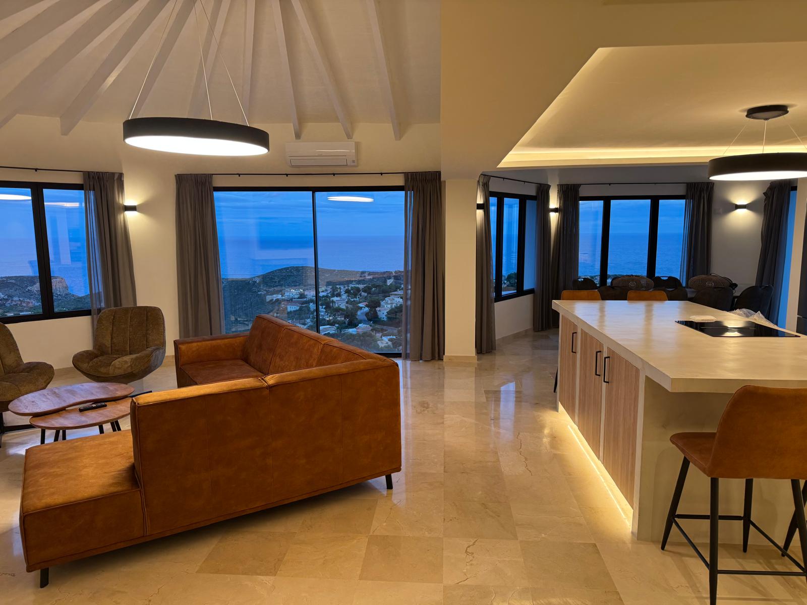 Open woonruimte met cognac lederen bank, kookeiland met LED-verlichting en grote ramen met panoramisch uitzicht op zee tijdens de schemering.
