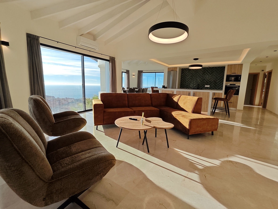 Open woonruimte met terracotta hoekbank, gewelfd balkenplafond en kamerhoge ramen met uitzicht op zee boven een gepolijste natuurstenen vloer.