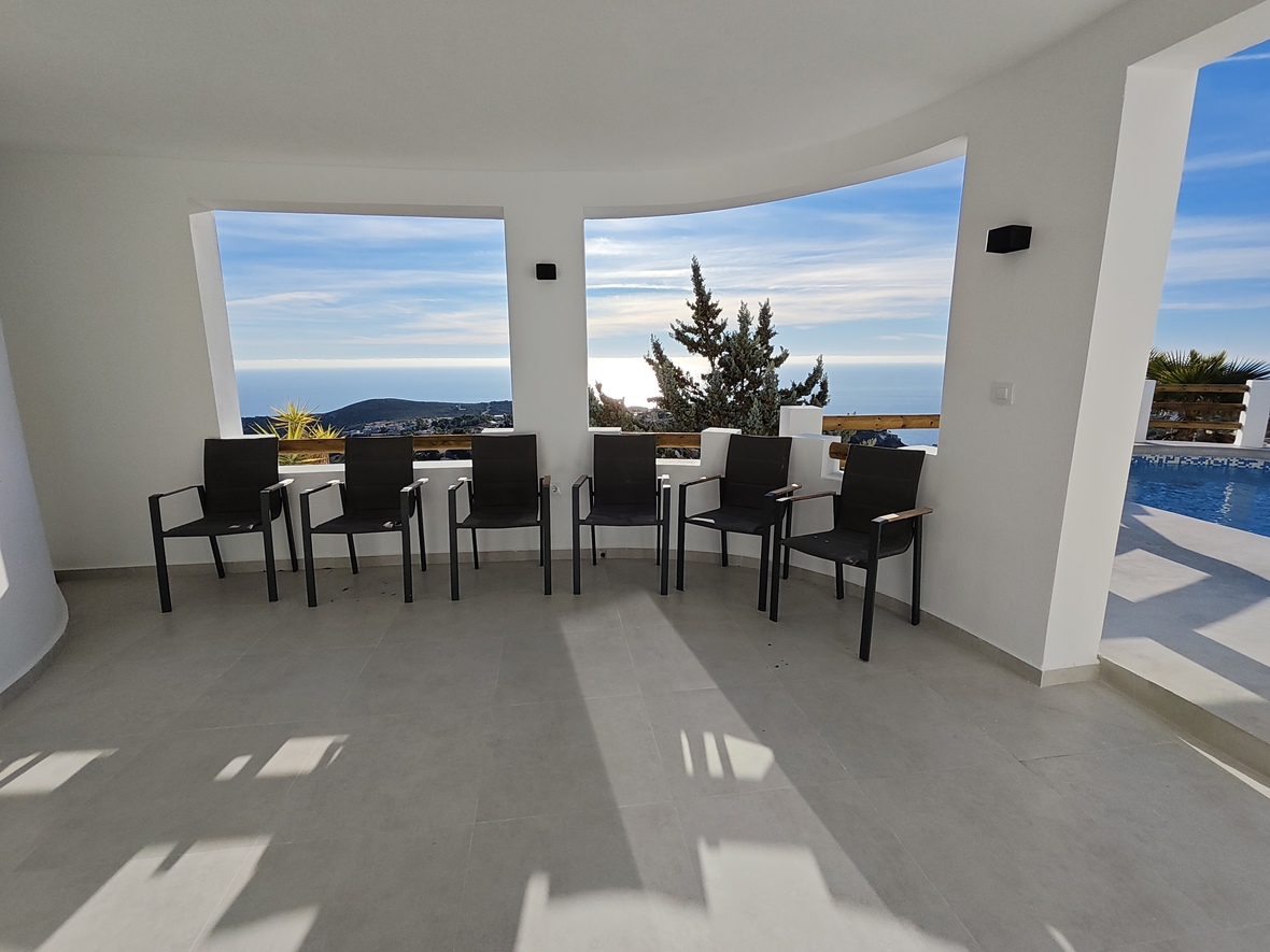 Overdekt terras met moderne zwarte fauteuils, lichtgrijze tegelvloer en grote muuropeningen met panoramisch uitzicht op de zee en het aangrenzende zwembad.