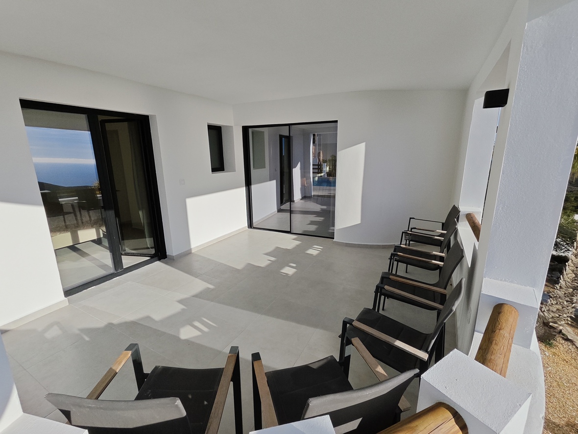 Overdekt terras met witte muren, lichtgrijze tegels en zwarte schuifdeuren. Voorzien van moderne zwarte stoelen met houten armleuningen en uitzicht op zee.