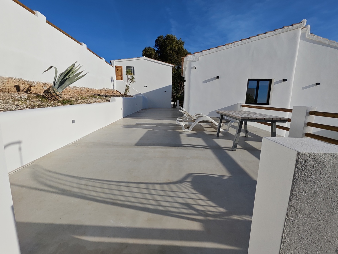 Ruim buitenterras met gepolijste betonvloer, witte ligbedden en een houten tafel, omgeven door witgekalkte mediterrane architectuur en een strakblauwe lucht.