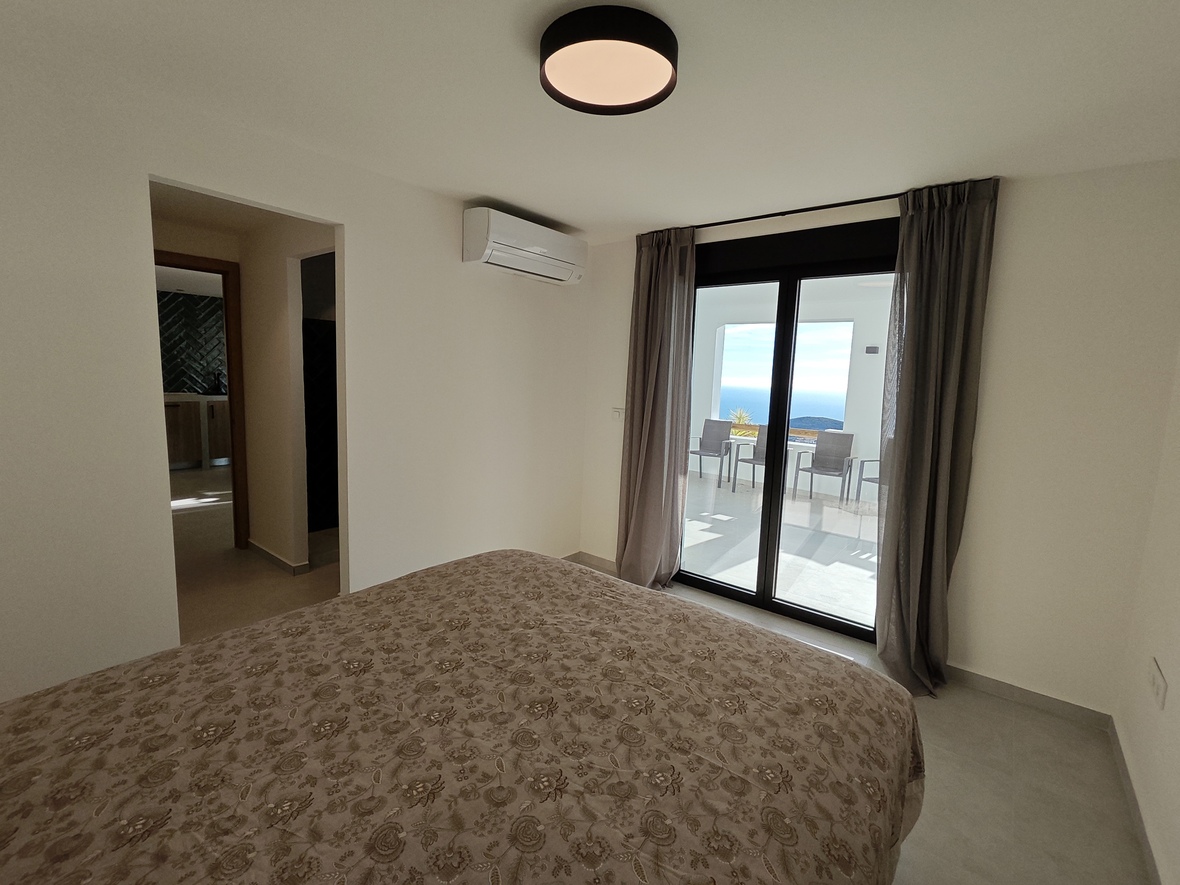 Slaapkamer met directe toegang tot een terras met uitzicht op zee. Voorzien van airconditioning, een neutraal decor en grote glazen schuifdeuren met grijze gordijnen.