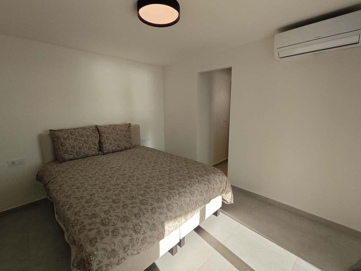 Slaapkamer met een tweepersoonsbed met dessin, split-unit airconditioning en een modern rond zwart armatuur op een lichte tegelvloer.