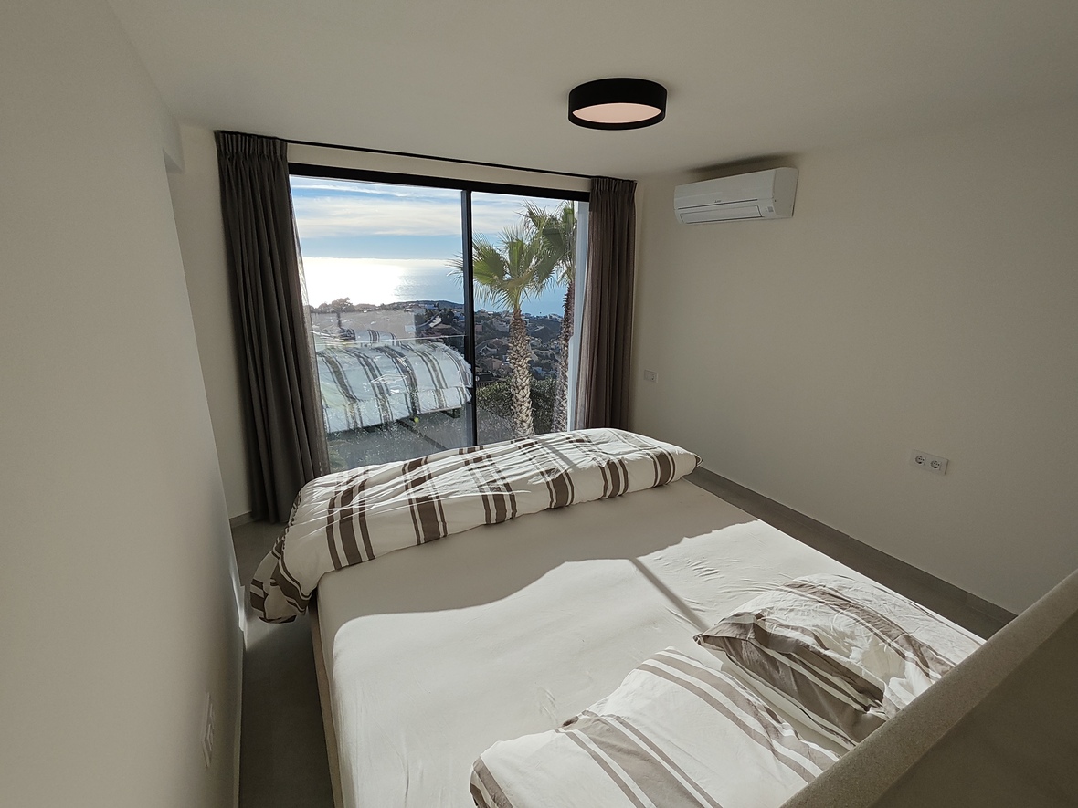 Slaapkamer met kamerhoge glazen schuifdeuren met uitzicht op zee en palmbomen. Voorzien van witte muren, gestreept beddengoed en wandgemonteerde airconditioning.