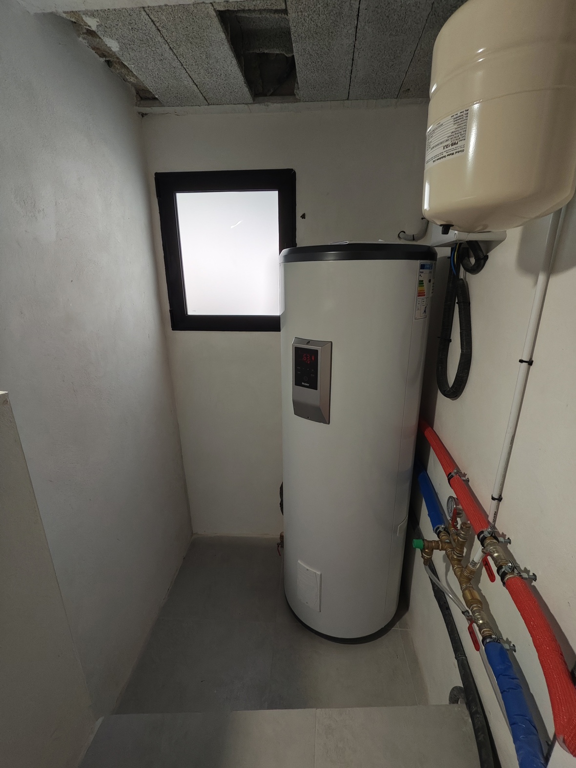 Technische ruimte met Haier warmtepompboiler, expansievat en geïsoleerde leidingen. Voorzien van een matglasraam en grijze tegelvloer.