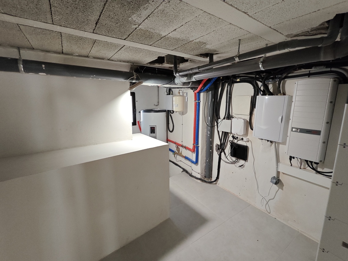 Technische ruimte met SolarEdge omvormer, elektrische panelen en warmwatersysteem met zichtbare PEX-leidingen en een betonnen plafond.