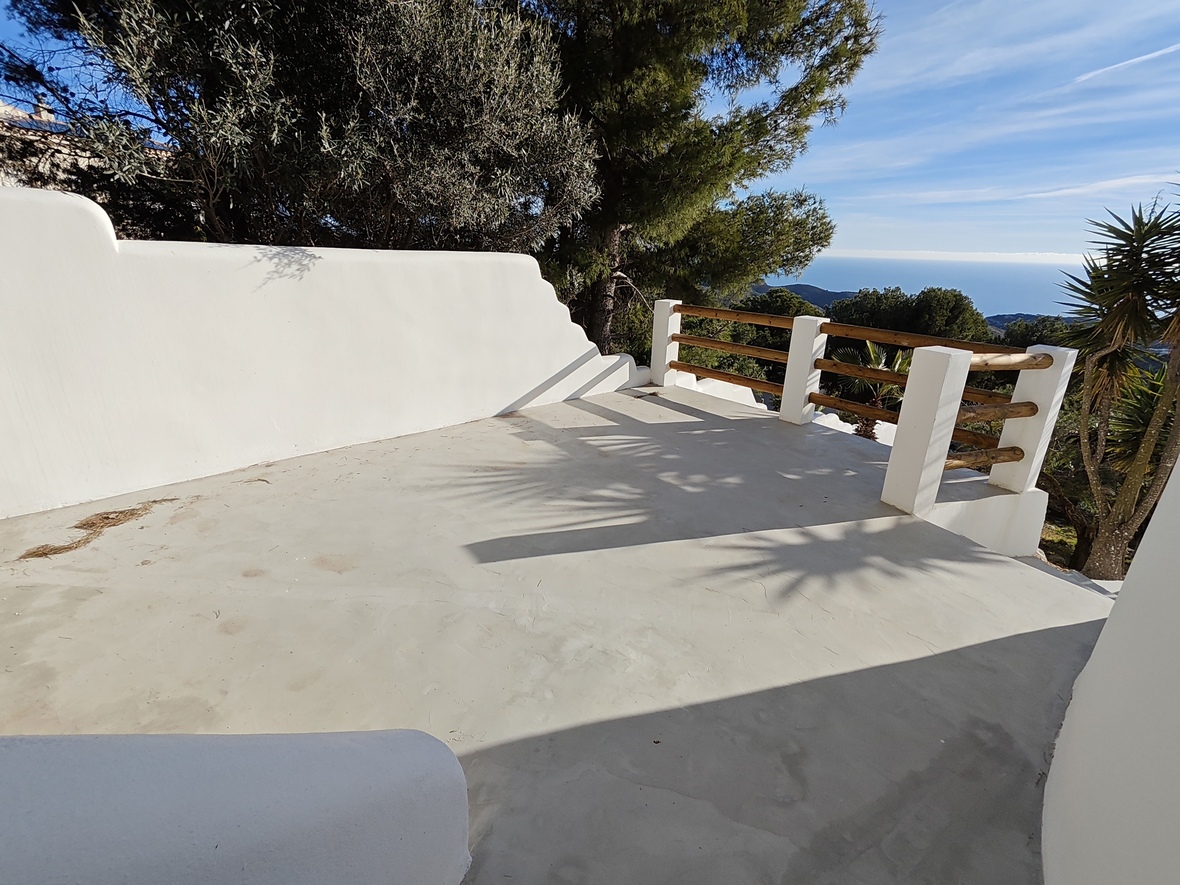 Verhoogd terras met gladde betonvloer, witgepleisterde muren en houten balustrade met panoramisch uitzicht op de Middellandse Zee en het omliggende dennenbos.