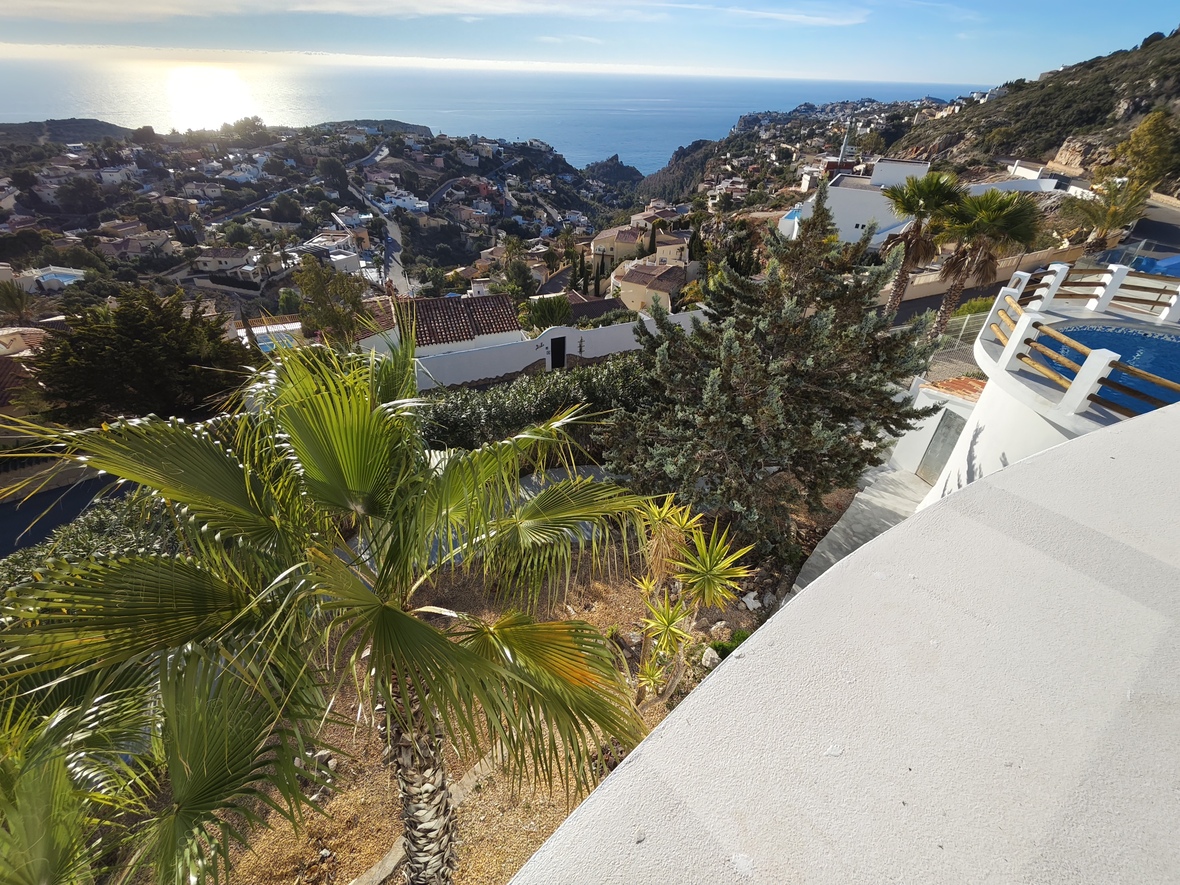 Verhoogd uitzicht vanaf een wit terras op een rond zwembad met houten reling, mediterrane tuinen en een panoramisch zicht op de glinsterende zee.