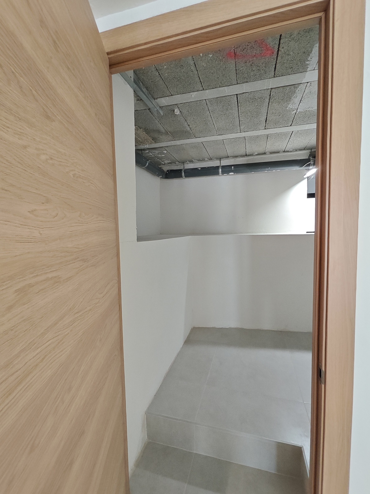 Zicht door een lichte houten deur op een berging of technische ruimte met grijze vloertegels en een onafgewerkt plafond met leidingen.