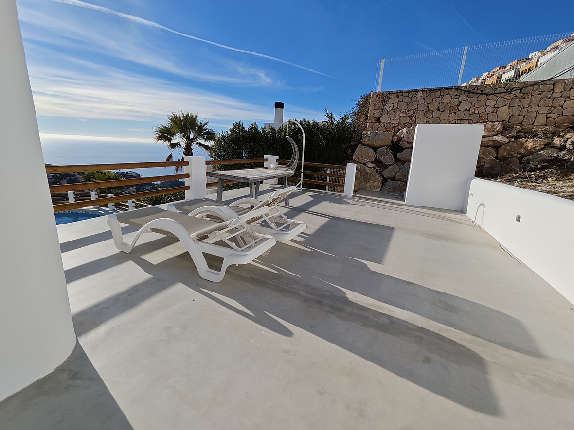 Zonneterras met witte ligbedden, houten tafel en hangstoel. Voorzien van panoramisch zeezicht, natuurstenen muren en een strakke betonvloer.