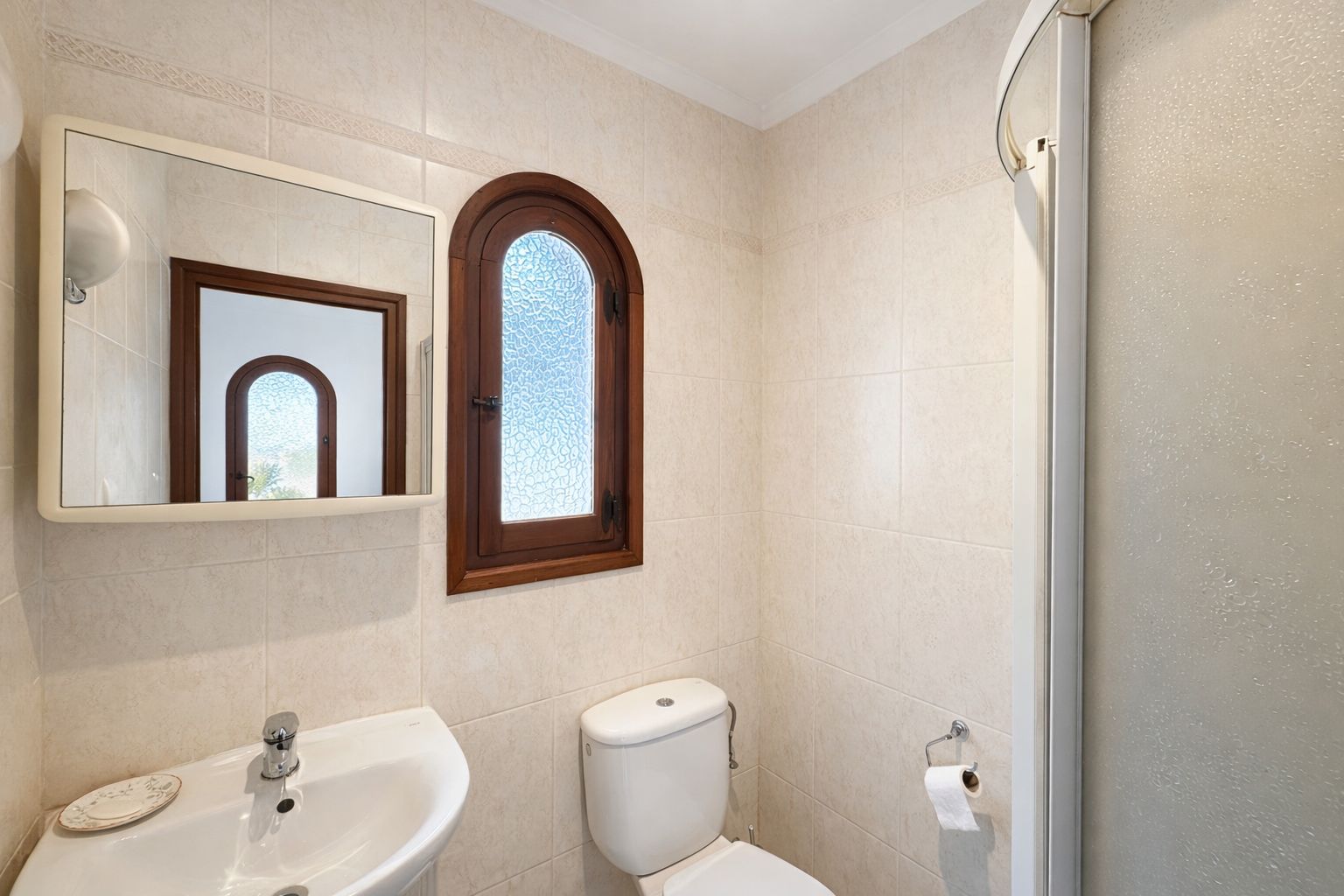 Badkamer met beige wandtegels, een houten boogvenster met getextureerd glas en een wit keramisch wastafelmeubel.