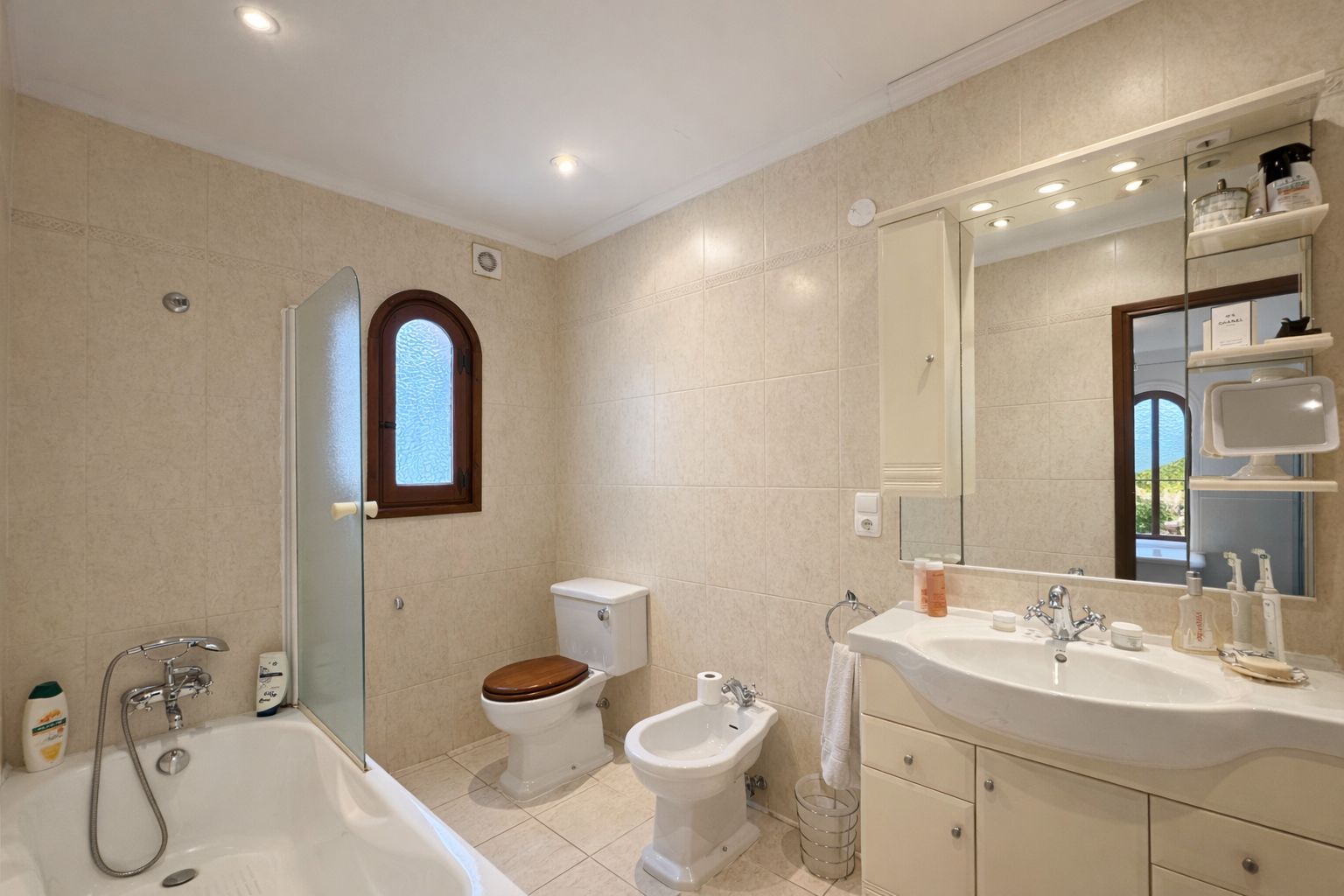 Badkamer met ligbad, glazen wand, bidet en toilet met houten zitting. Voorzien van een wastafelmeubel, grote spiegel met verlichting en een houten boograam.