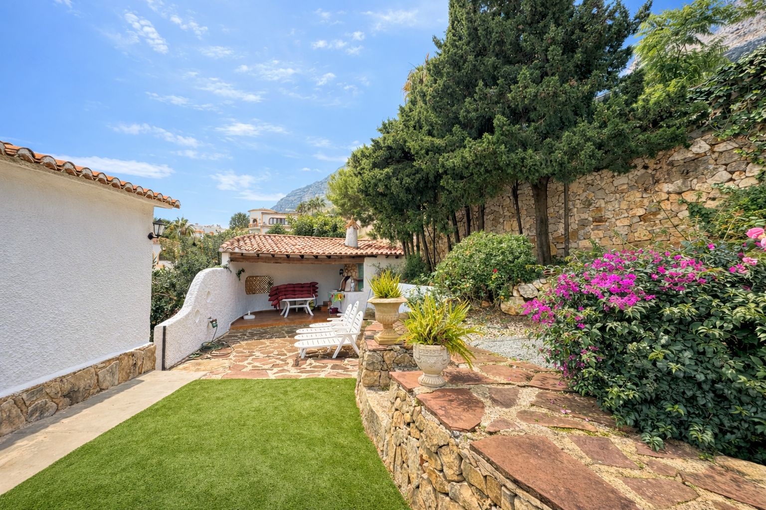 Buitenterras met natuurstenen bestrating, kunstgras en een overdekte veranda met terracotta pannen. Natuurstenen muren en mediterrane beplanting sieren de tuin.