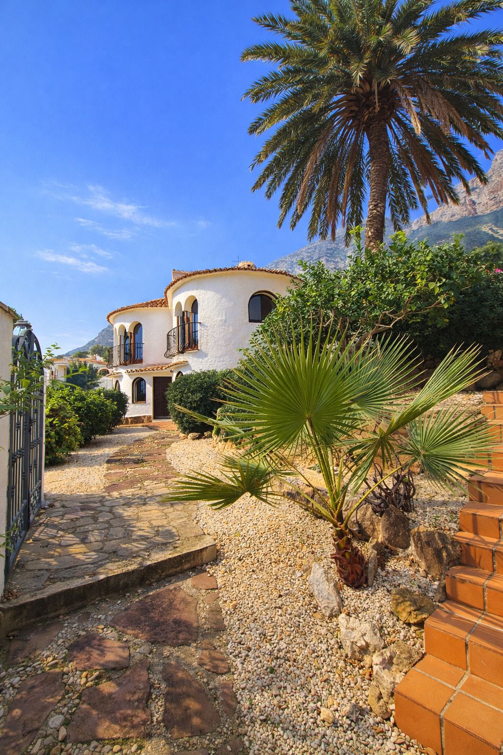 Exterieur van een witte mediterrane villa met ronde vormen, boogvensters en een stenen pad in een tuin met palmbomen en uitzicht op de bergen.
