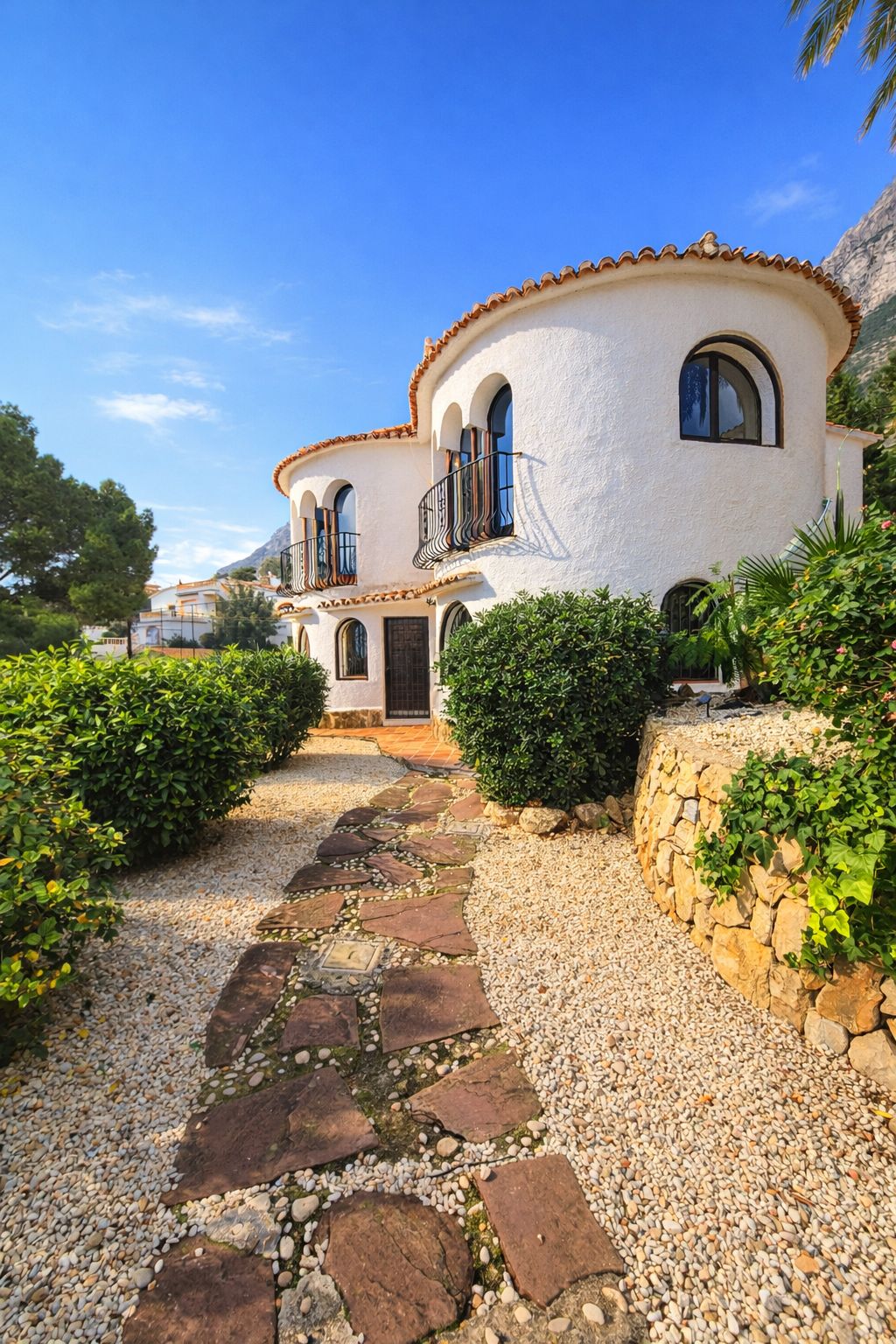 Mediterrane villa met witte ronde muren, boogvensters en smeedijzeren balkons. Een pad van natuursteen loopt door de grindtuin met groene beplanting.