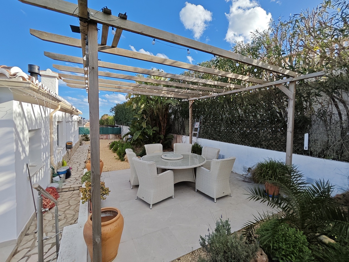 Buiten eethoek met een ronde glazen tafel en zes witte rotan stoelen onder een houten pergola met lichtsnoeren en mediterrane beplanting.