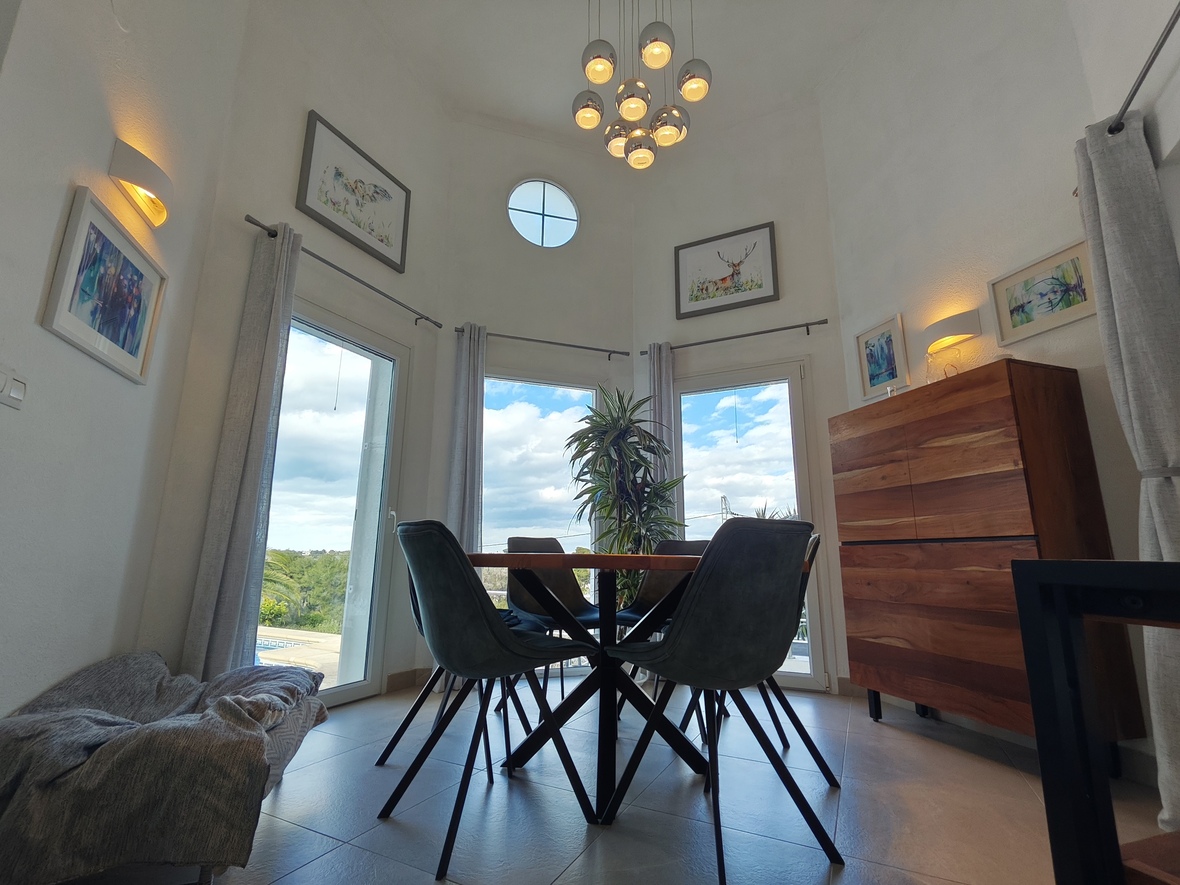 Eethoek met hoge plafonds, een moderne chromen hanglamp en een rond venster. Voorzien van een houten dressoir en grote ramen die uitkijken op de omliggende tuin.