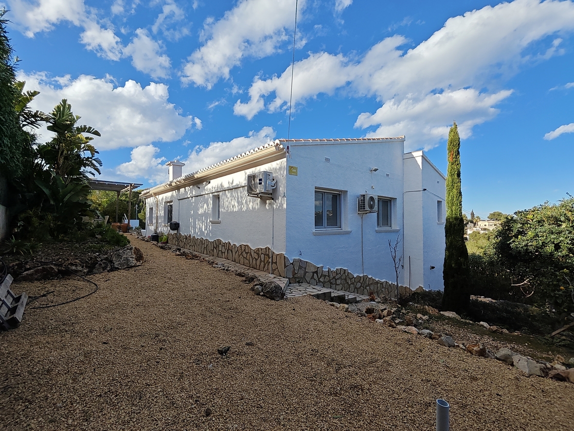 Exterieur van de villa met witte muren en een natuurstenen plint, grenzend aan een grindpad en mediterrane beplanting onder een helderblauwe lucht.