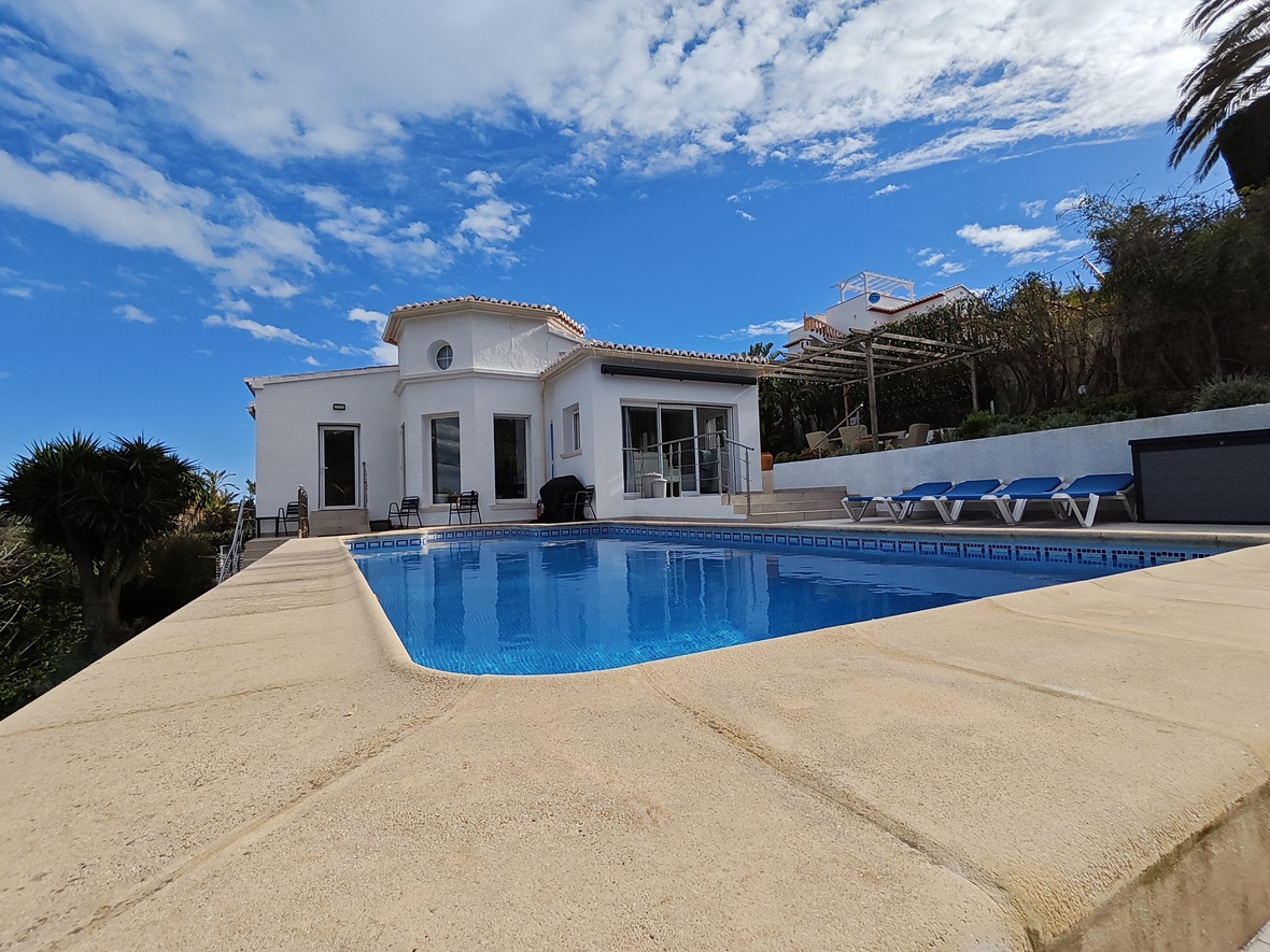 Exterieur van een witte villa met privézwembad, stenen terras, blauwe ligbedden en mediterrane architectuur tegen een heldere lucht.