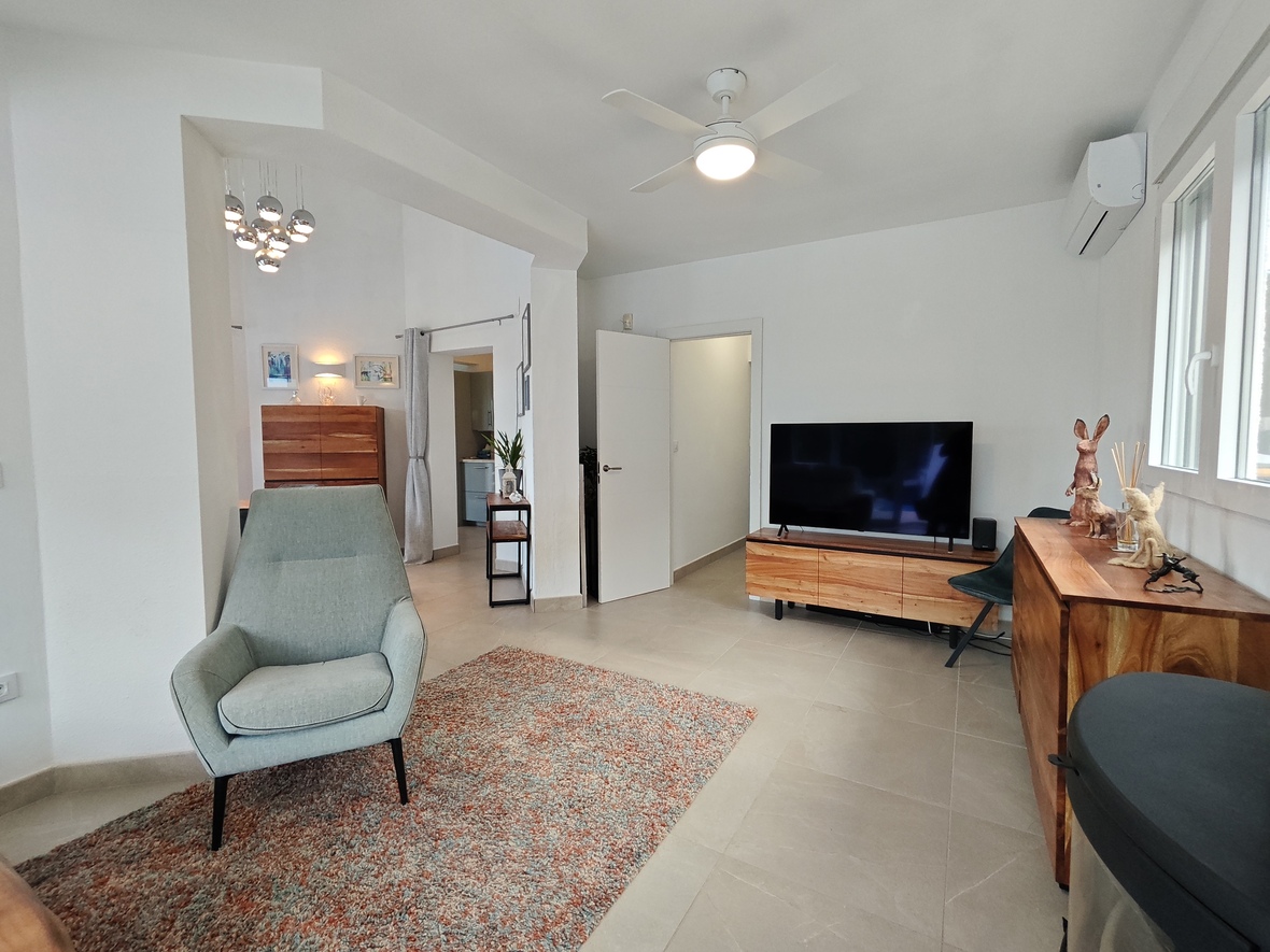 Lichte woonkamer met een moderne fauteuil, houten mediameubel en flatscreen-tv. Voorzien van een plafondventilator, airconditioning en een neutrale tegelvloer.