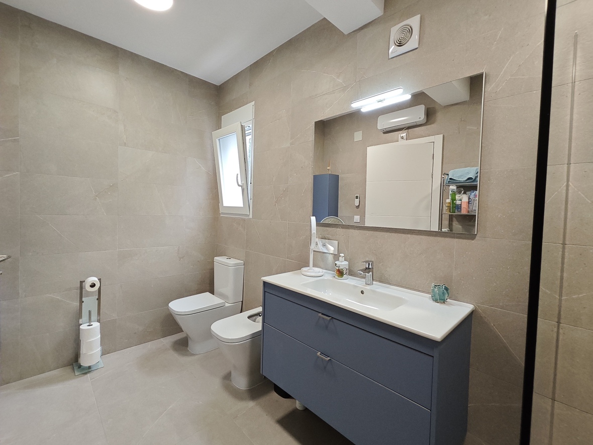 Moderne badkamer met beige tegels in steenlook, een blauw zwevend badkamermeubel met geïntegreerde wastafel en een toilet-bidet combinatie onder een verlichte spiegel.