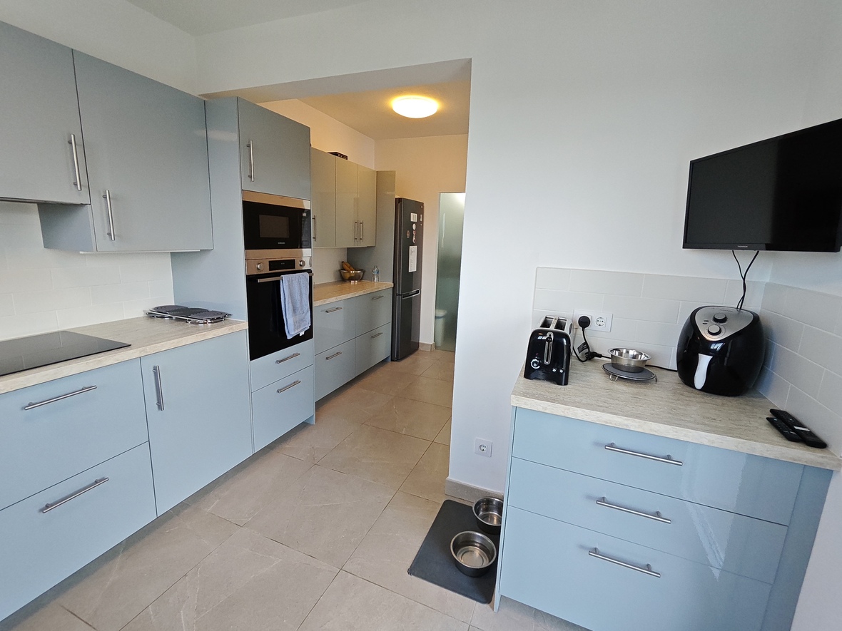 Moderne keuken met lichtblauwe kasten, geïntegreerde apparatuur en werkbladen met steeneffect. Voorzien van een wandtelevisie en grijze vloertegels.