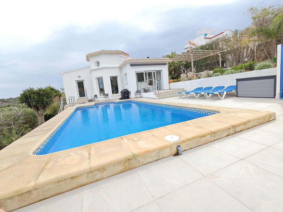 Privé rechthoekig zwembad met blauwe tegels en stenen rand, omgeven door een licht betegeld terras met ligbedden en een witte mediterrane villa.