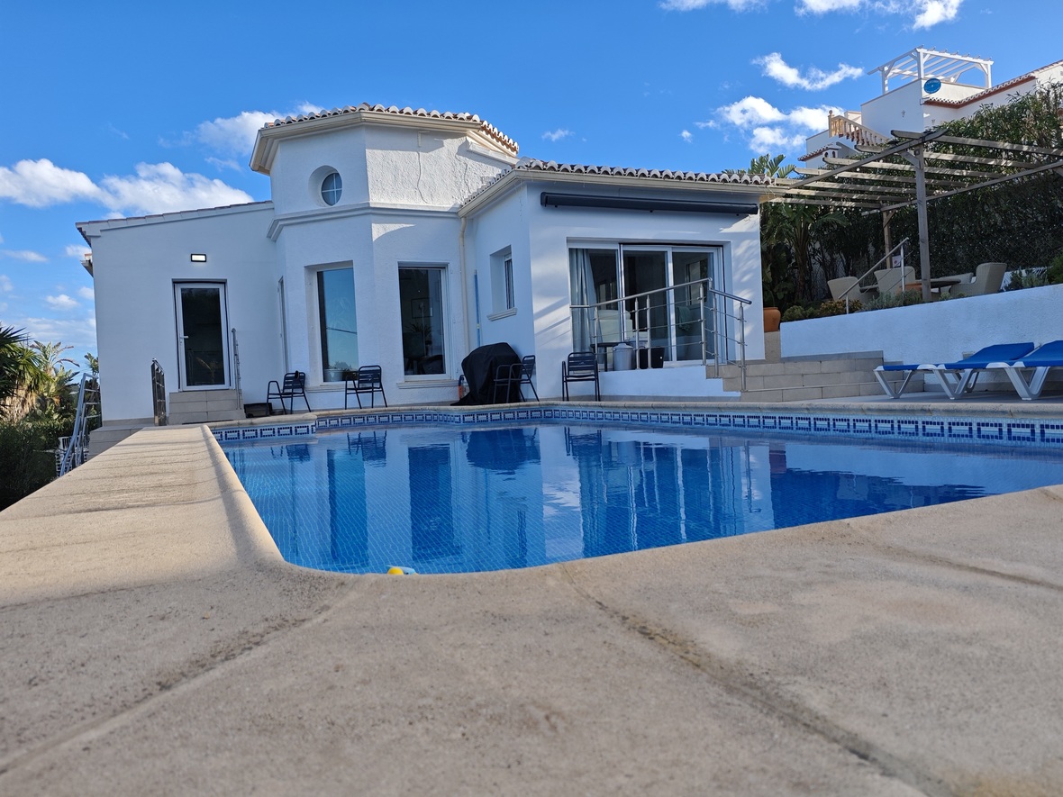 Privézwembad met blauwe mozaïektegels en stenen terras, gelegen bij een witte mediterrane villa met een achthoekige toren en grote glazen schuifdeuren.