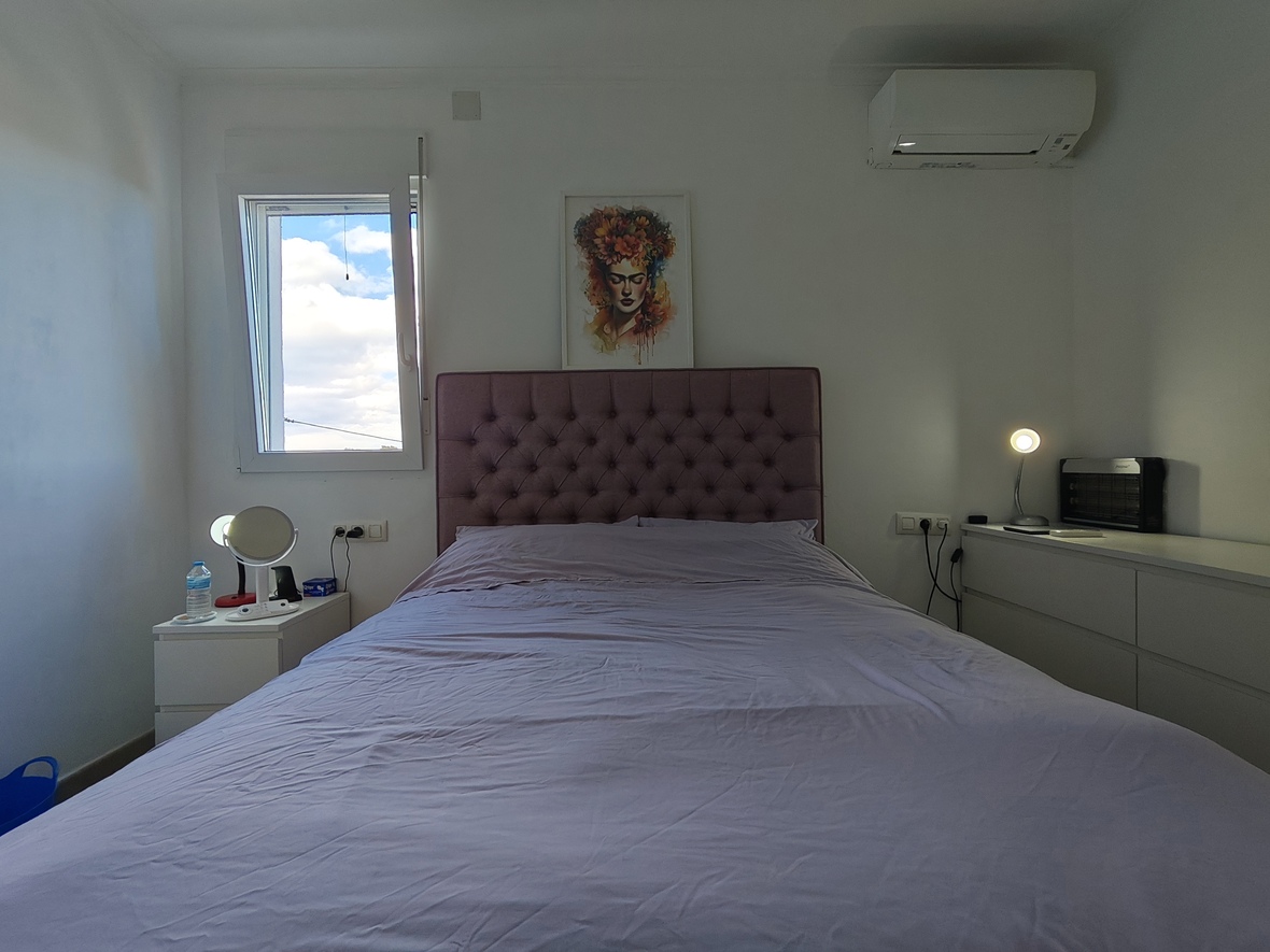 Slaapkamer met een mauve gecapitonneerd hoofdbord, witte minimalistische meubels en airconditioning. Een portret hangt boven het bed bij een raam met daglicht.