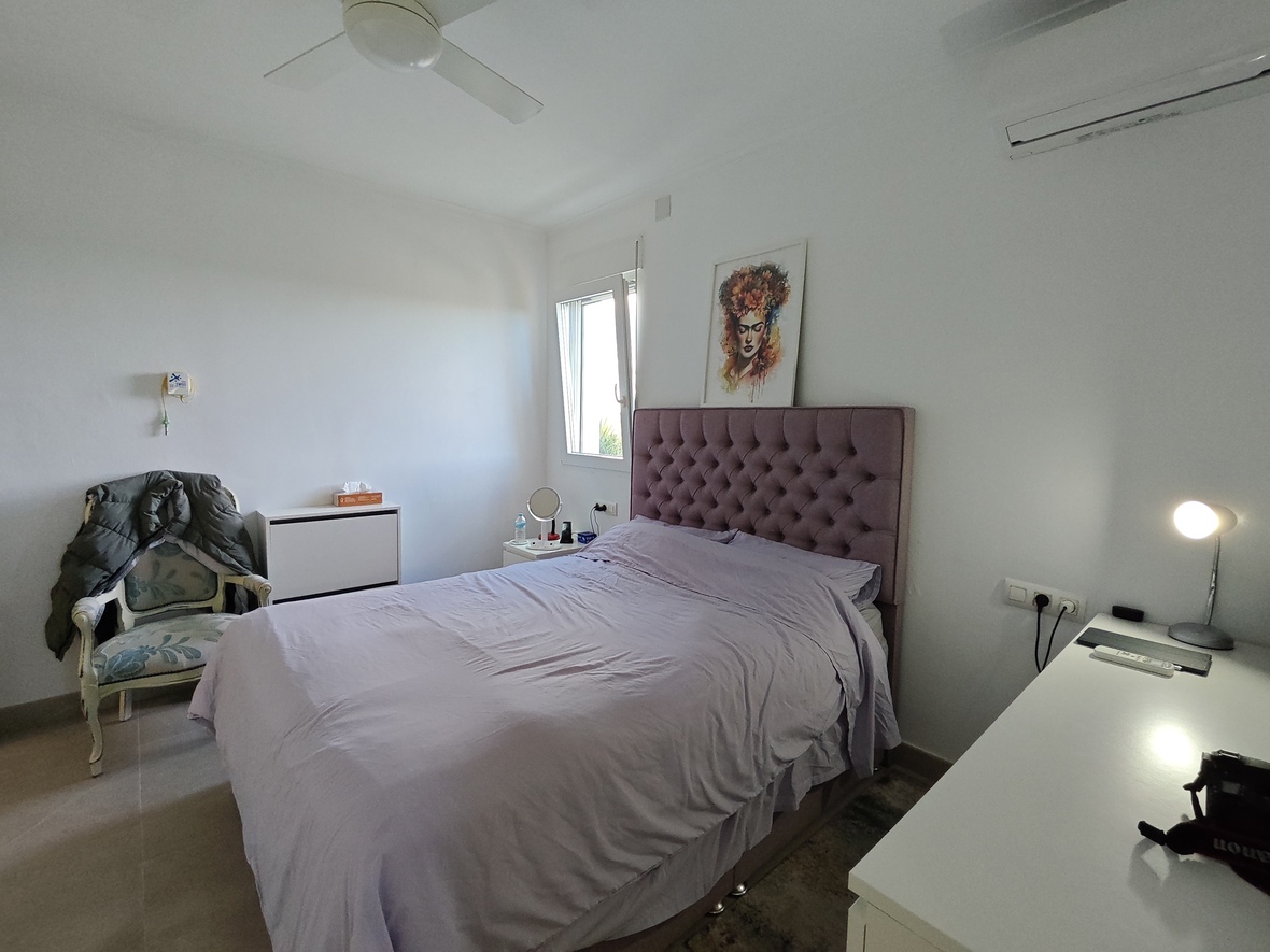 Slaapkamer met een mauve gecapitonneerd hoofdbord, witte muren, plafondventilator en een praktische werkplek met bureau en natuurlijk licht.