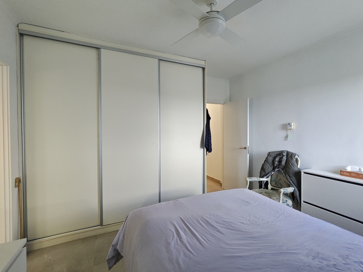 Slaapkamer voorzien van een grote driedeursschuifkast, een witte plafondventilator en een klassieke fauteuil naast een moderne ladekast.