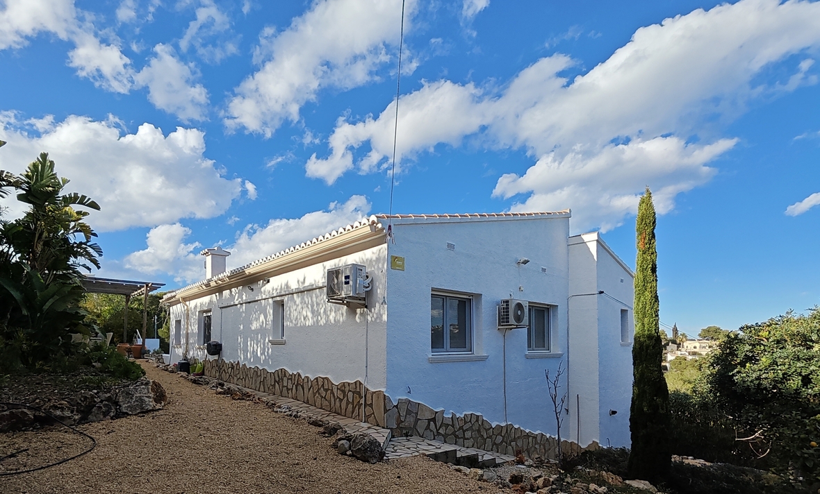 Witte buitengevel met natuurstenen plint, grindpad en mediterrane beplanting onder een helderblauwe lucht.