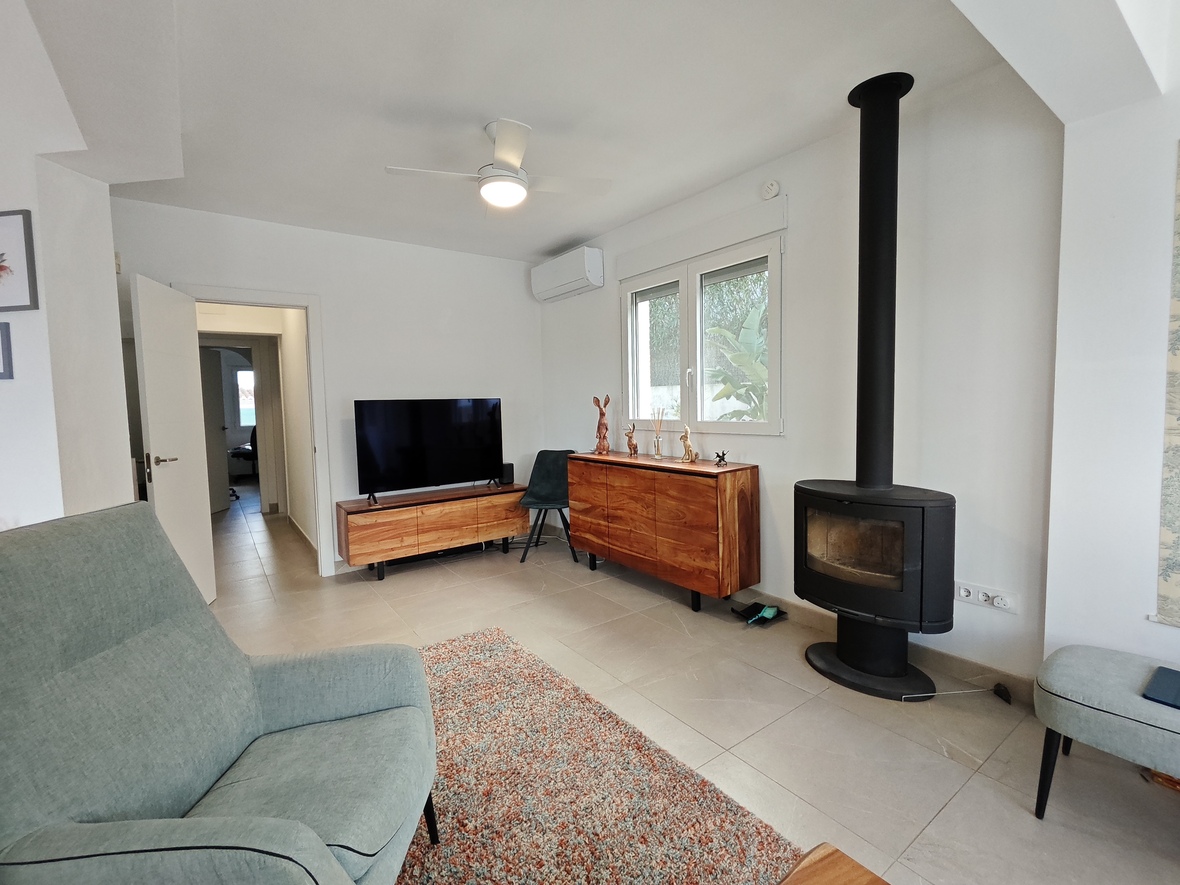 Woonkamer met moderne houtkachel, massief houten dressoirs en een lichte tegelvloer. Voorzien van airconditioning, een plafondventilator en uitzicht op de tuin.