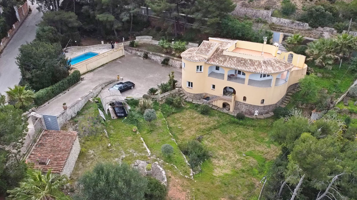 Luchtfoto van de villa met gebogen gevel en stenen plint, voorzien van een ruime oprit, privézwembad en een terrasvormige tuin omgeven door pijnbomen.