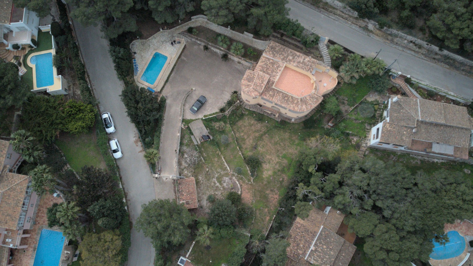 Luchtfoto van het perceel met de indeling van de villa, de ruime verharde oprit, het privézwembad en de terrasvormige tuin omgeven door mediterrane pijnbomen.