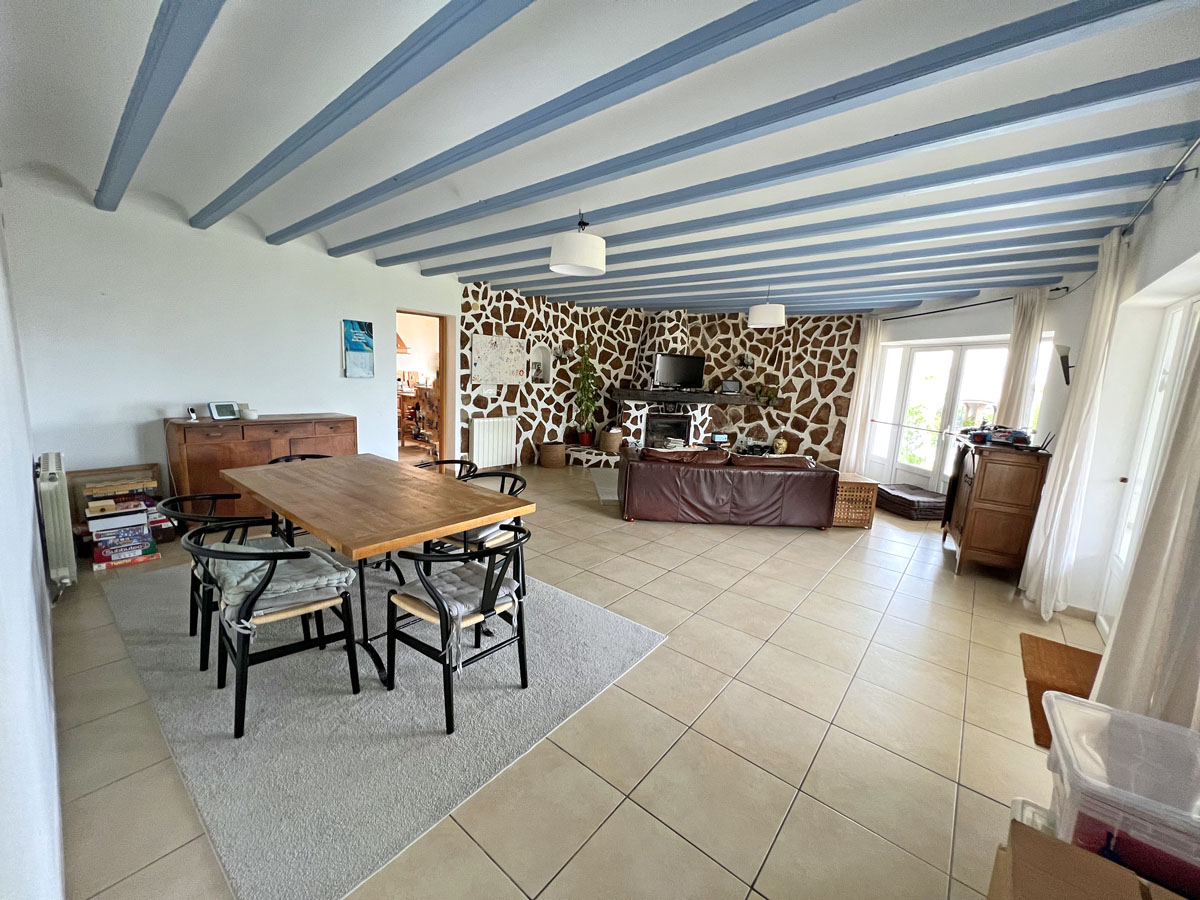 Open woon- en eetkamer met blauw geverfde plafondbalken, een natuurstenen accentmuur met open haard en grote keramische vloertegels.