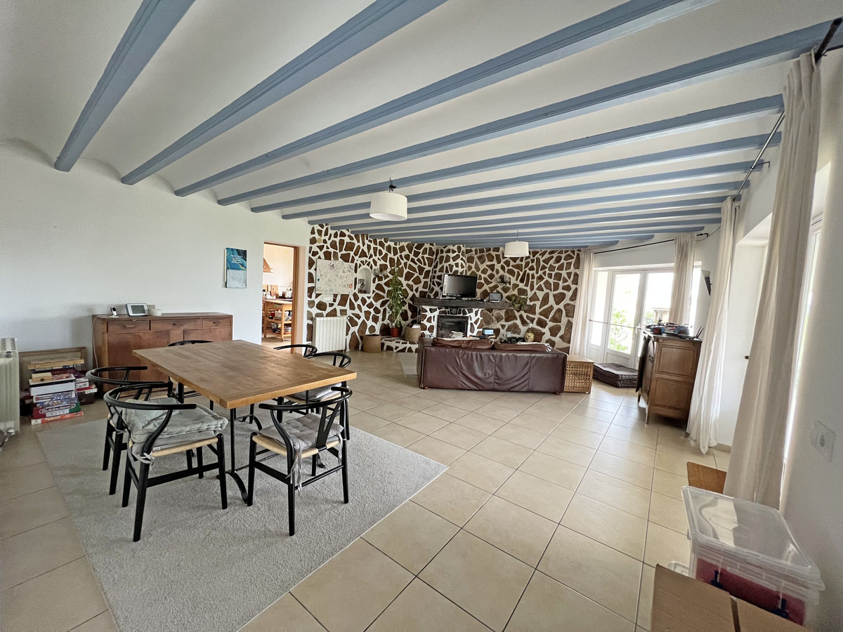 Open woon- en eetkamer met lichtblauwe houten plafondbalken, een decoratieve natuurstenen wand met haard en een neutrale keramische tegelvloer.
