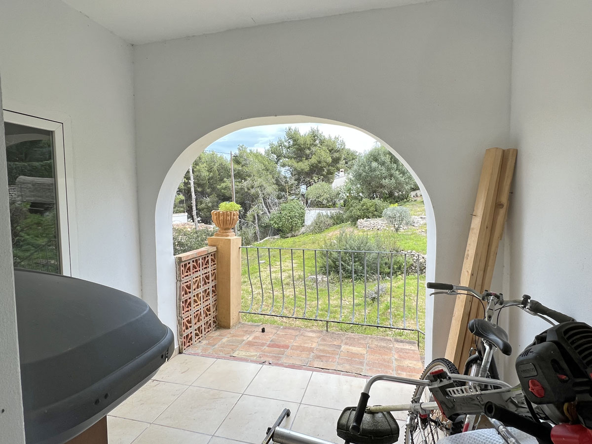 Overdekte veranda met een karakteristieke boog en uitzicht op de mediterrane tuin en pijnbomen, voorzien van een tegelvloer en metalen balustrade.