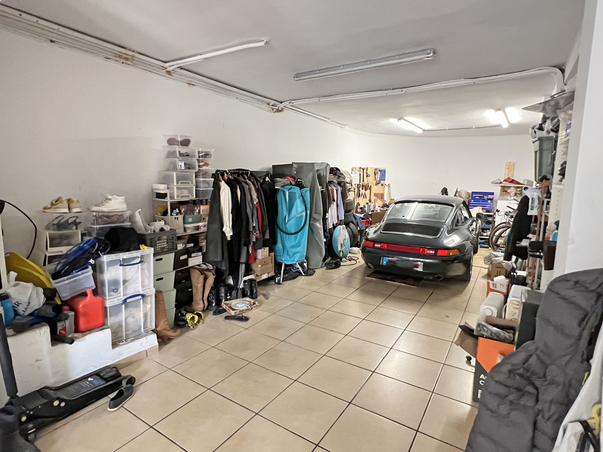 Ruime garage met tegelvloer, tl-verlichting en voldoende ruimte voor voertuigen en georganiseerde opslag- of werkplaatsruimte.