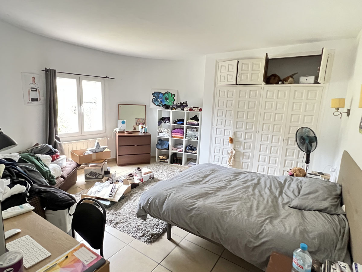 Slaapkamer met een grote witte inbouwkast met paneeldeuren, zijraam voor natuurlijk licht en een tegelvloer. De ruimte biedt plaats voor een bureau en opslag.