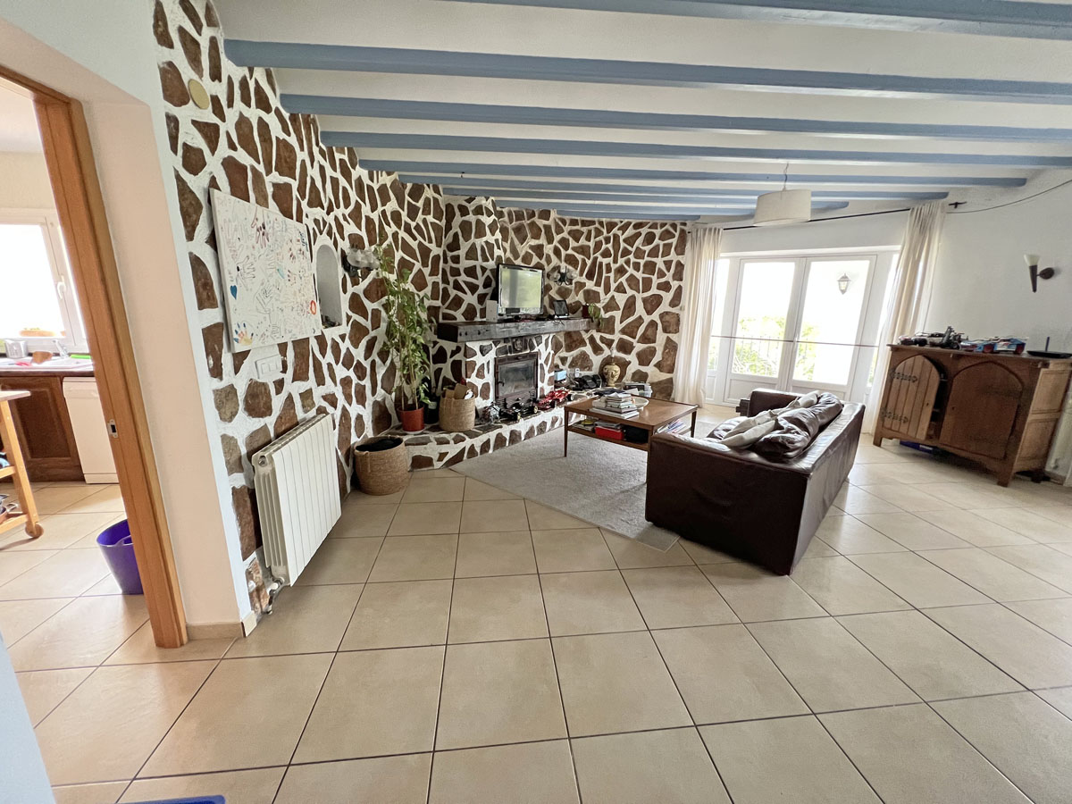 Woonkamer met een karakteristieke natuurstenen wand, ingebouwde haard, blauwe plafondbalken en directe toegang tot het terras.