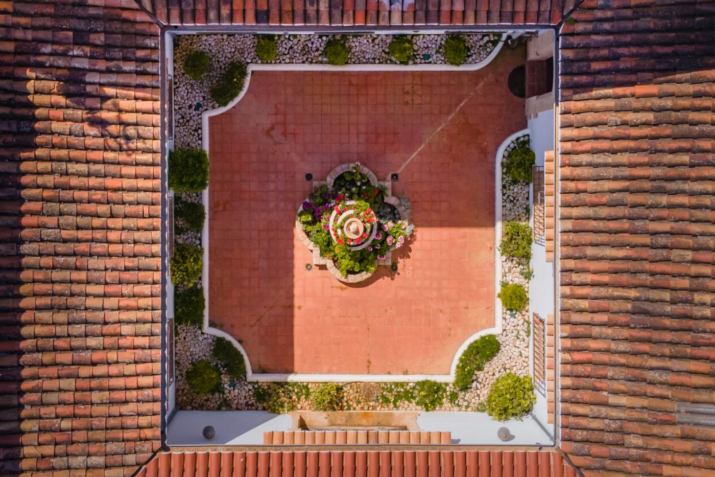 Bovenaanzicht van de centrale binnenplaats met terracotta tegels, een bloemrijke fontein en omliggende borders, omringd door traditionele pannendaken.