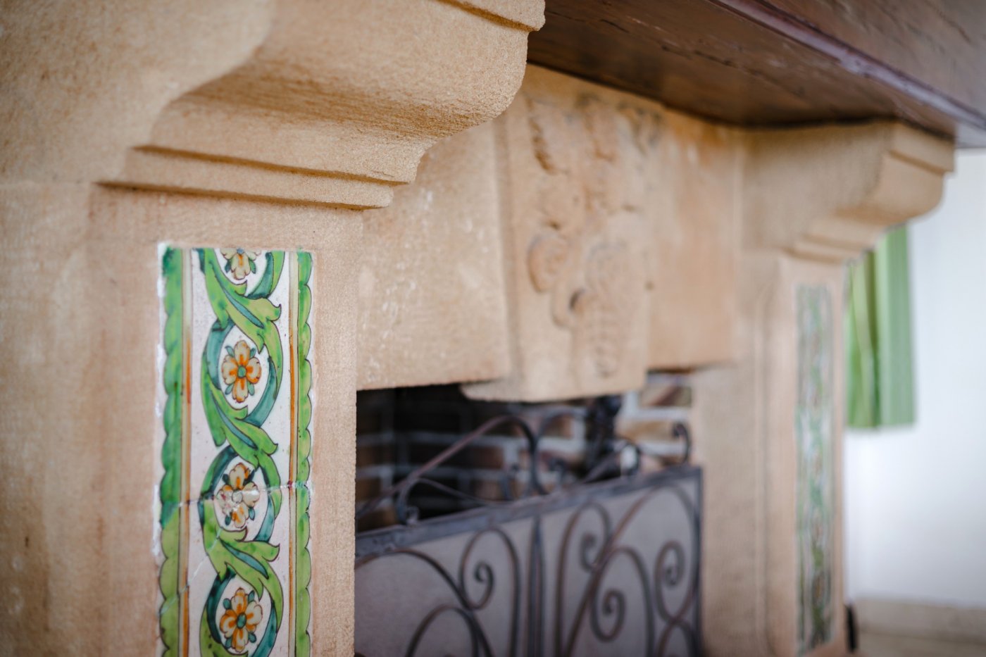 Close-up van een traditionele open haard van Tosca-steen met handbeschilderde keramische tegels, een decoratief reliëf en een smeedijzeren scherm.