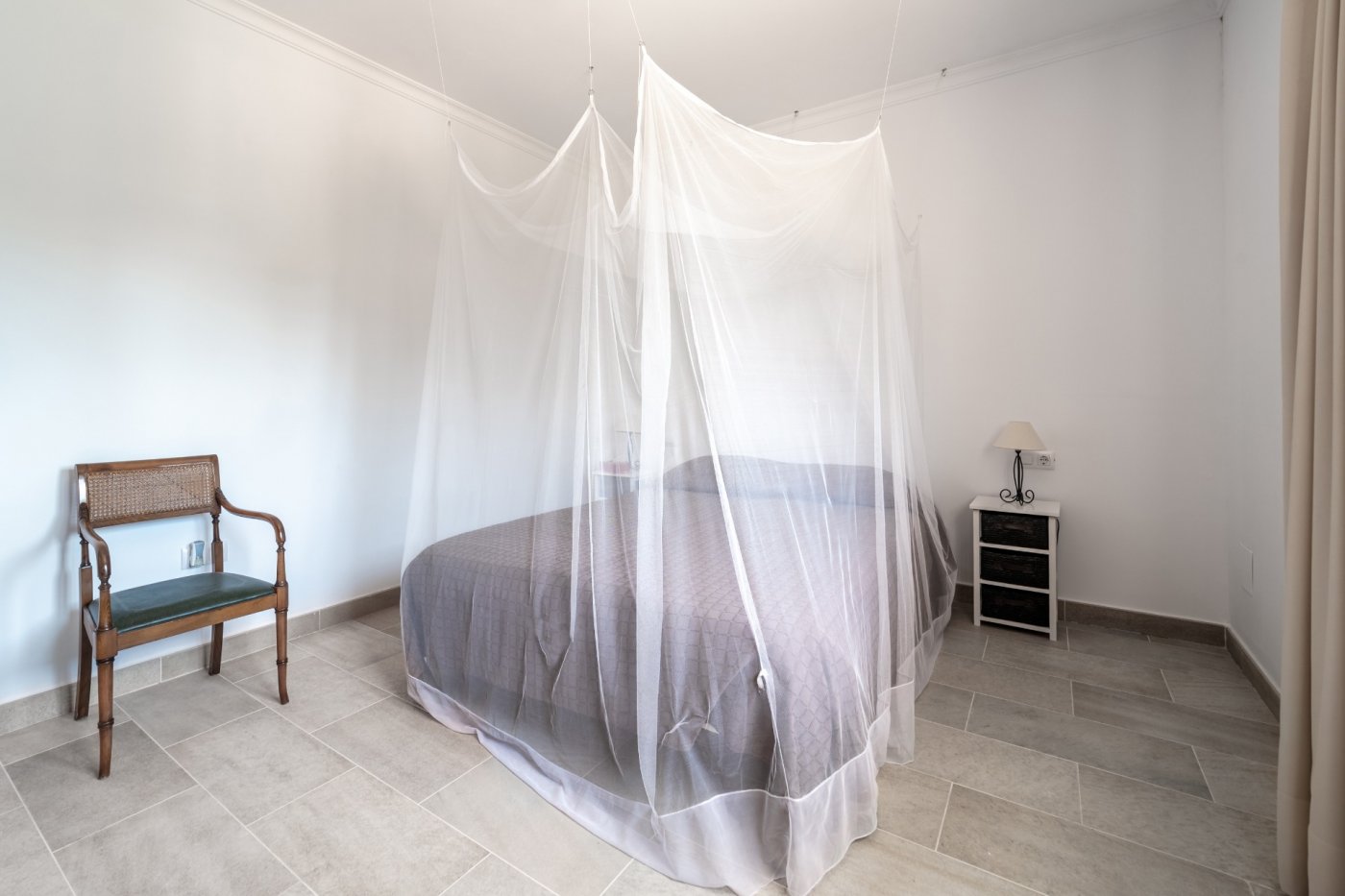 Lichte slaapkamer met een vloer van keramische tegels, een tweepersoonsbed met muggennet en klassiek meubilair tegen strakke witte wanden.