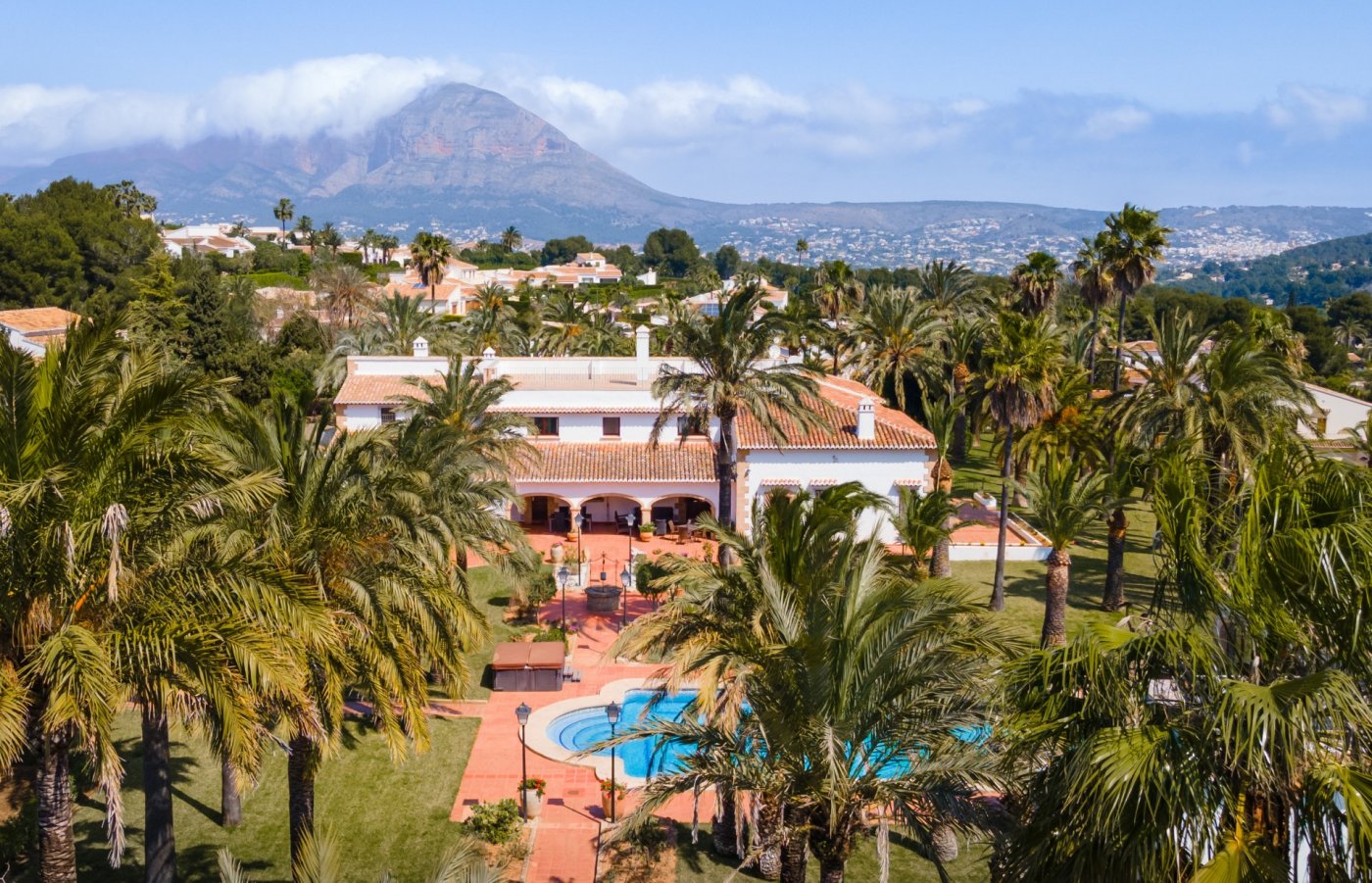 Luchtfoto van de mediterrane villa met een groot zwembad, uitgestrekte palmentuinen en panoramisch uitzicht op de berg Montgó op de achtergrond.