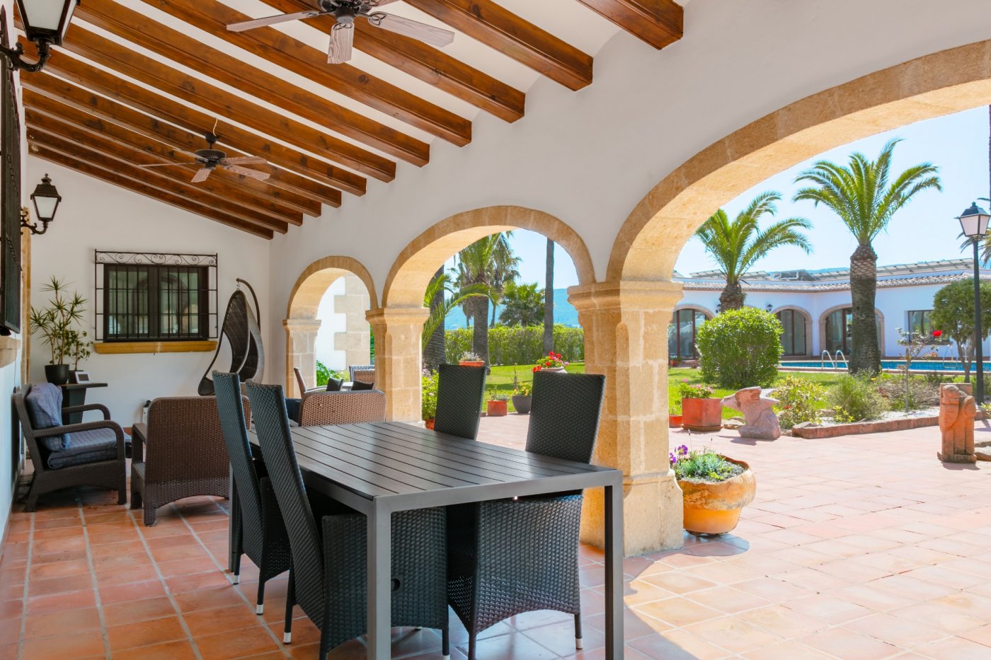 Ruim overdekt terras met traditionele Tosca stenen bogen, houten balkenplafonds en een eethoek met uitzicht op de mediterrane tuin met palmbomen.