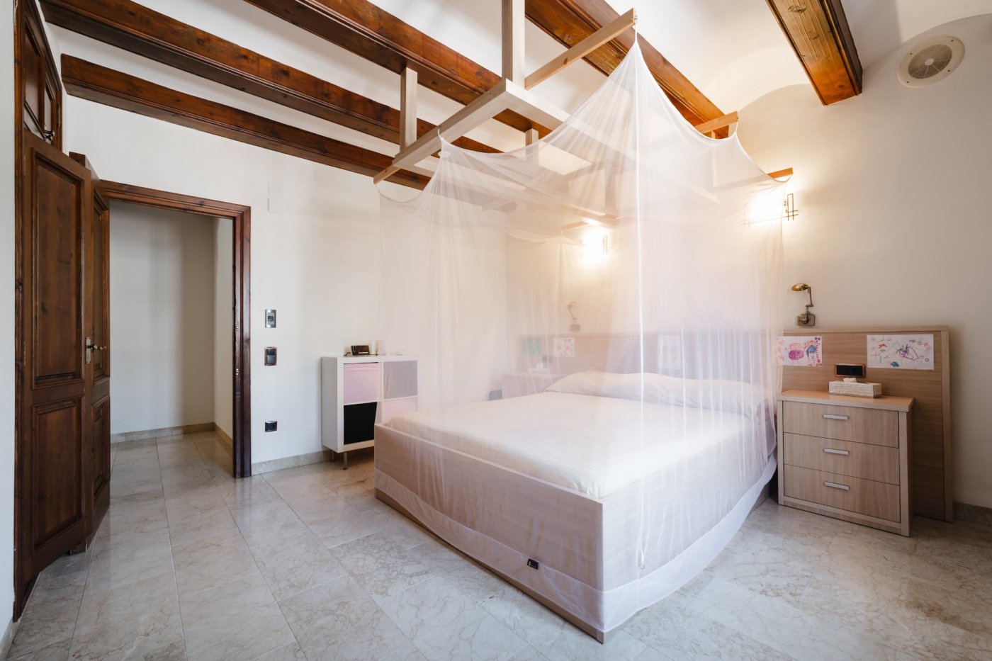 Slaapkamer met donkere houten balkenplafonds, een bed met muskietennet en een lichte natuurstenen vloer. Voorzien van massief houten deuren en wandverlichting.