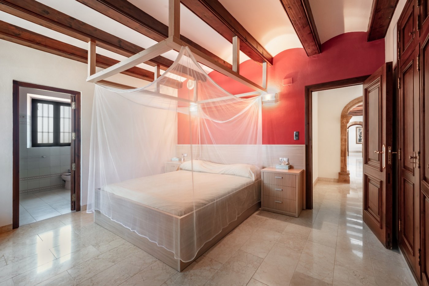 Slaapkamer met houten balken, een rode accentmuur en hemelbed. Inclusief marmeren vloer, badkamer en suite en zicht op de karakteristieke stenen bogen.