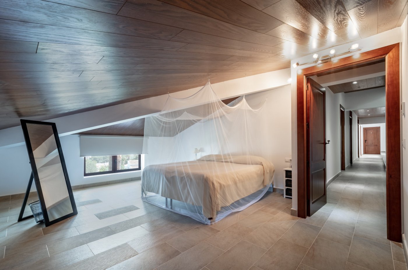 Slaapkamer onder het schuine dak met houten plafondafwerking, bed met klamboe en plavuizen vloer. De ruimte is voorzien van een passpiegel en grenst aan een gang.