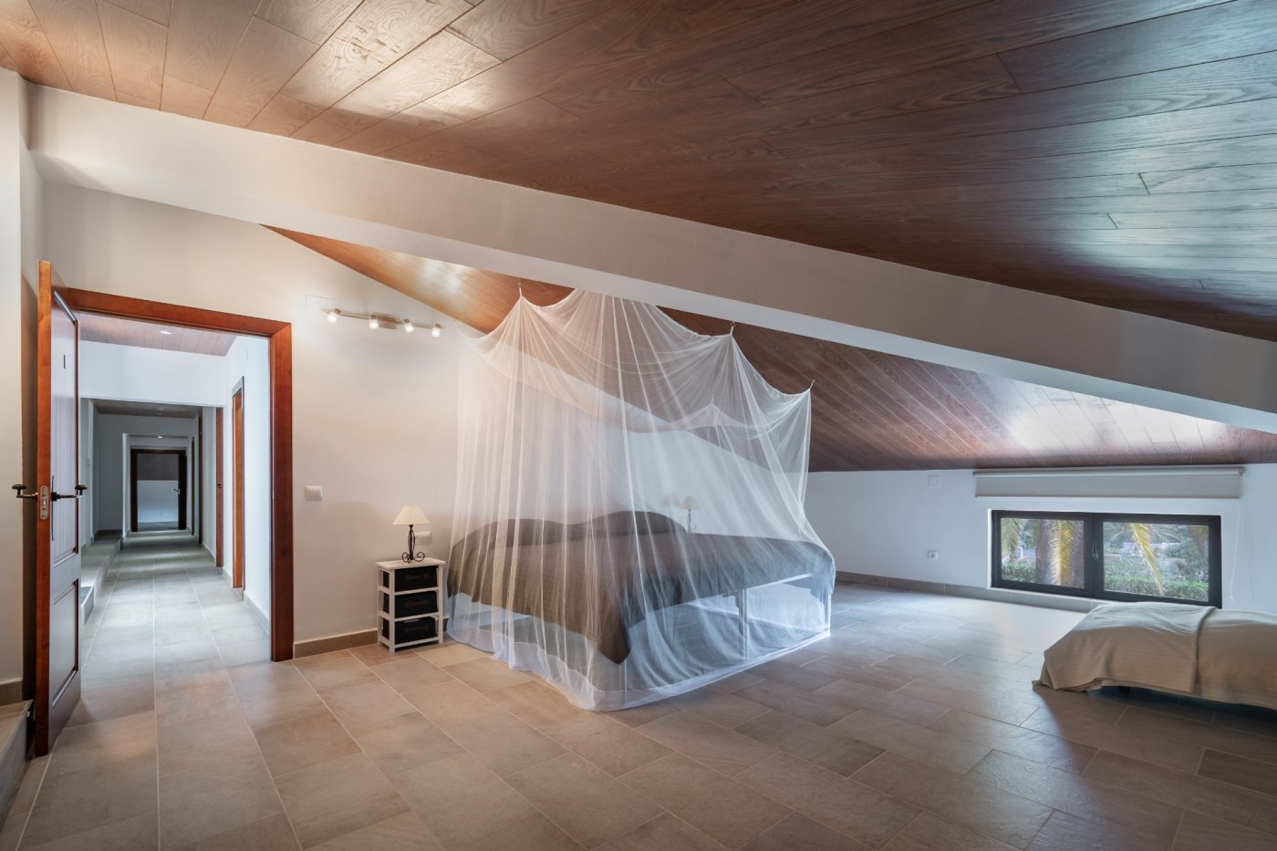 Slaapkamer op de bovenverdieping met schuin houten plafond, tegelvloer en een bed met muskietennet. Een laag raam kijkt uit op de tuin en palmbomen.